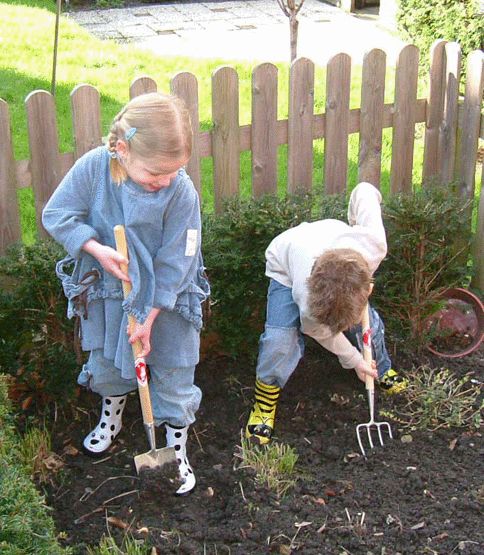 Zwei Kinder im Garten beim Spielen mit Gartengeräten. Ein Mädchen mit einer Hacke und ein Junge mit einer Forke, beide in Regenstiefeln und Gartenkleidung.